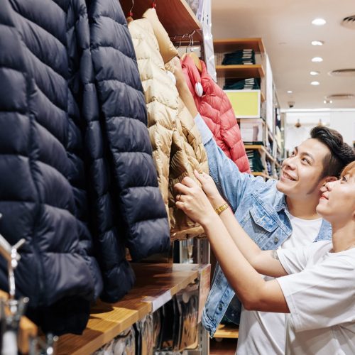 Positive young Asian couple reaching for warm winter jacket on hanger in store